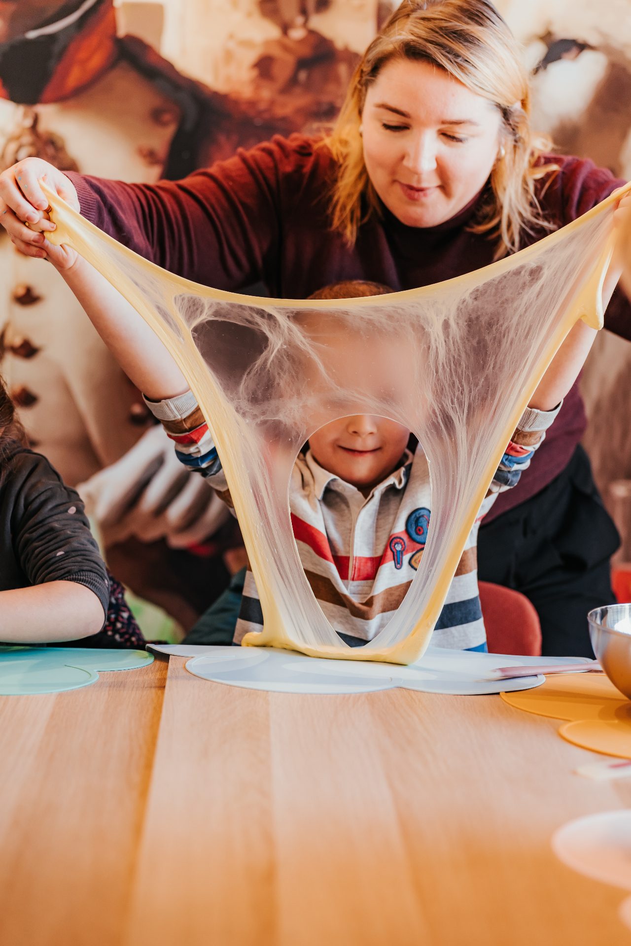 Atelier Slime Art Studio — animatrice étirant un slime jaune translucide en toile d'araignée au-dessus d'un enfant souriant à table