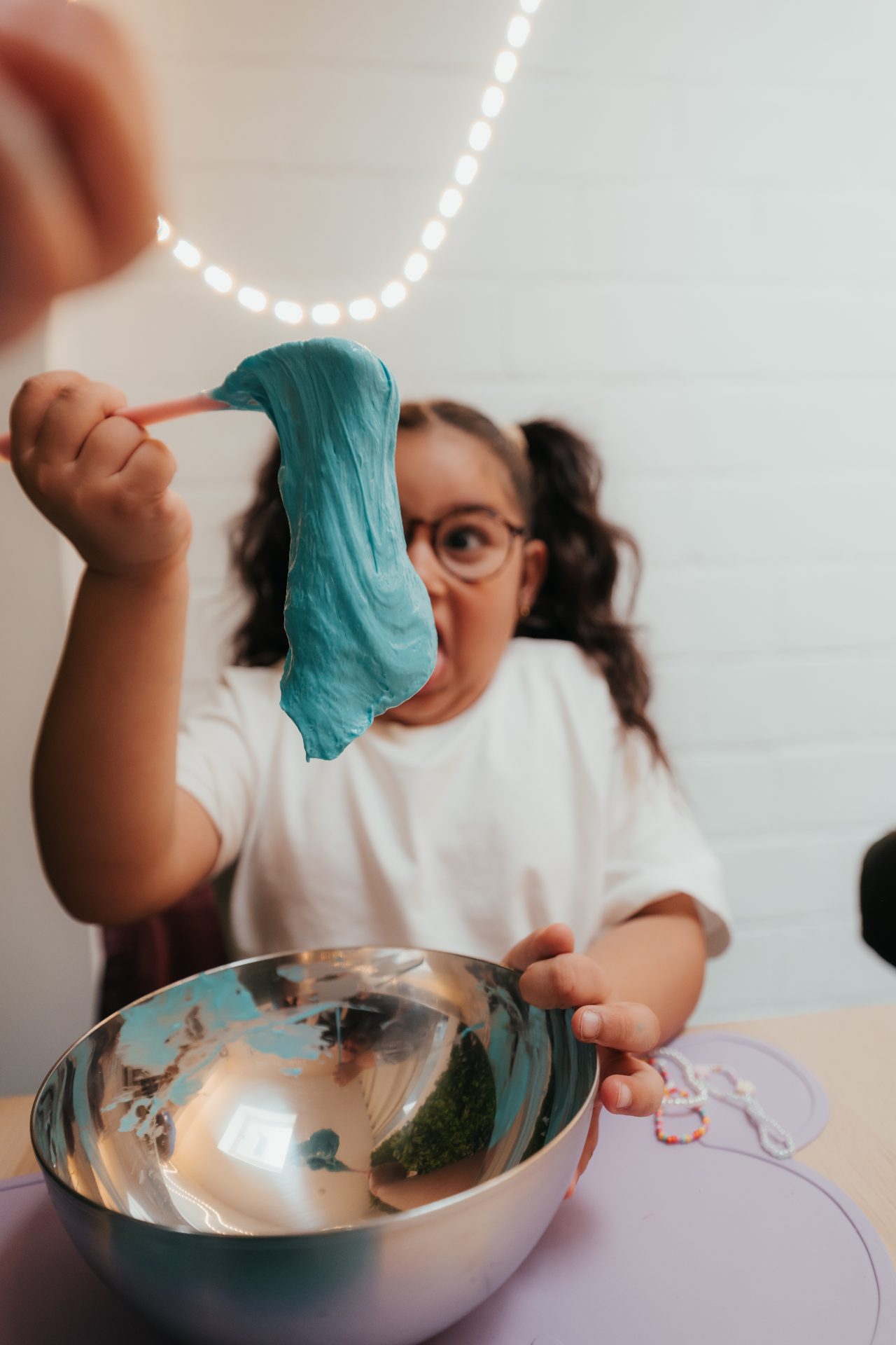Atelier Slime Art Studio — petite fille émerveillée étirant sa slime bleue devant un bol en inox, sous une guirlande lumineuse