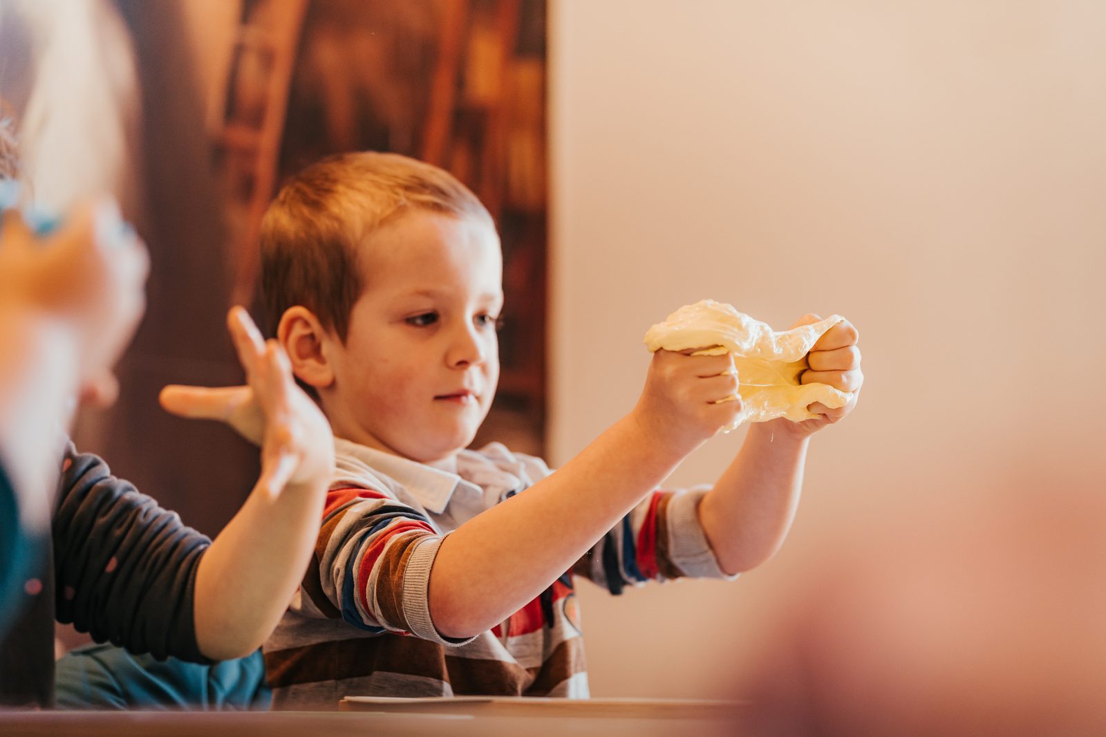 Atelier Slime Art Studio — petit garçon concentré étirant une slime jaune pastel entre ses mains dans une salle chaleureuse