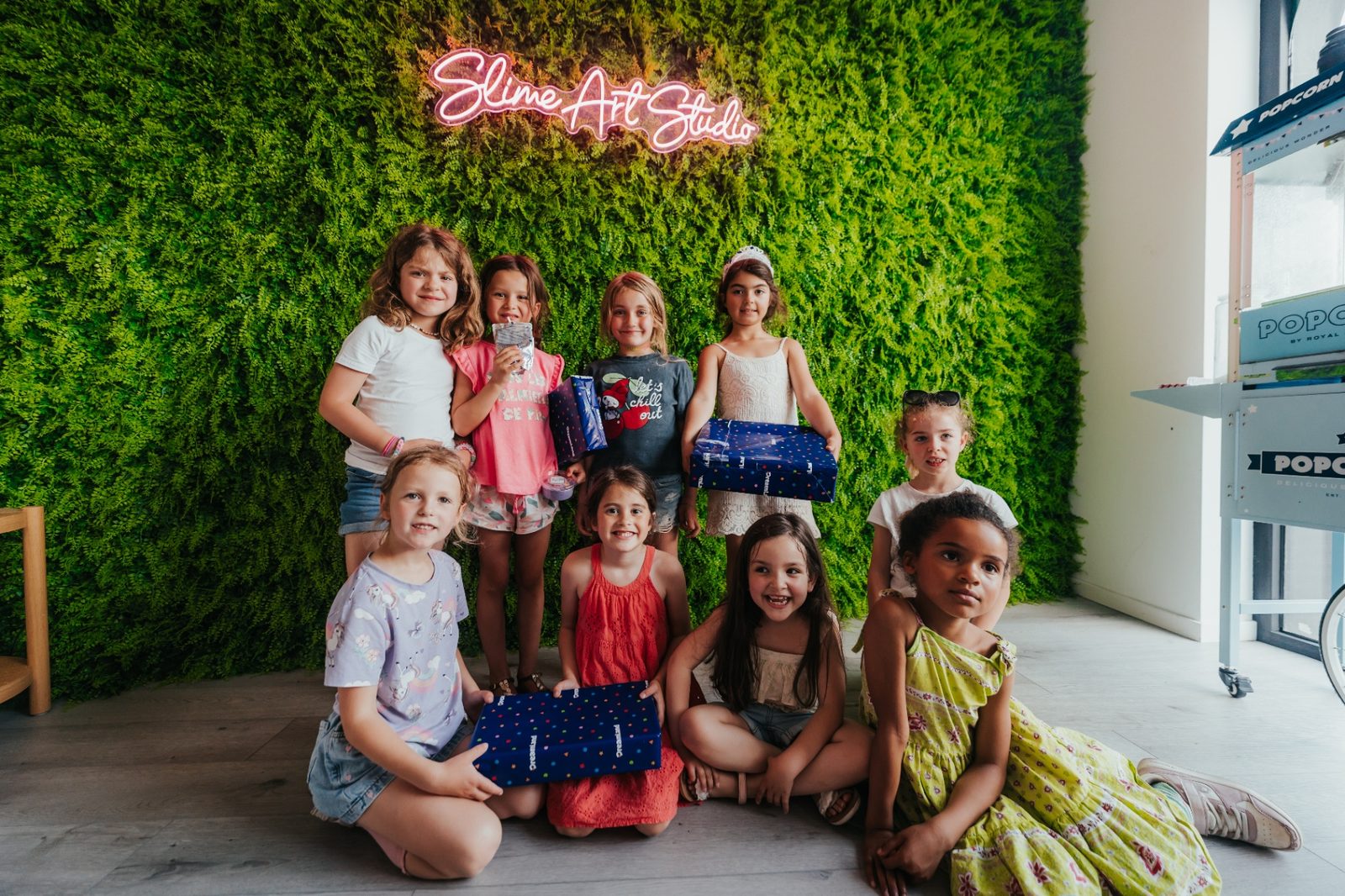 Groupe d'enfants souriant avec leurs cadeaux devant le mur végétal et l'enseigne néon Slime Art Studio après un anniversaire