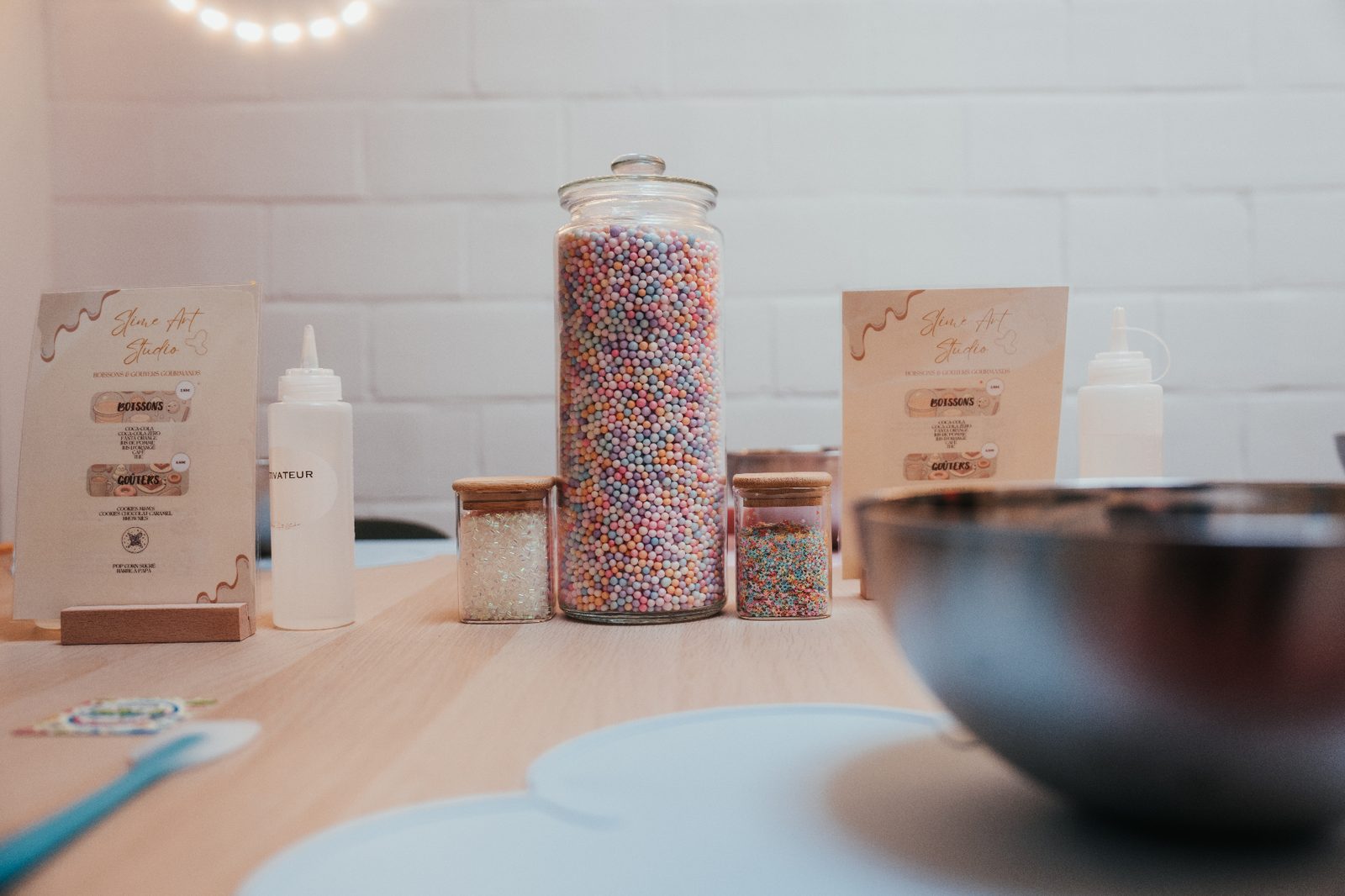 Atelier slime à Mons — bocal de perles pastel multicolores, paillettes et menu Slime Art Studio sur table en bois clair