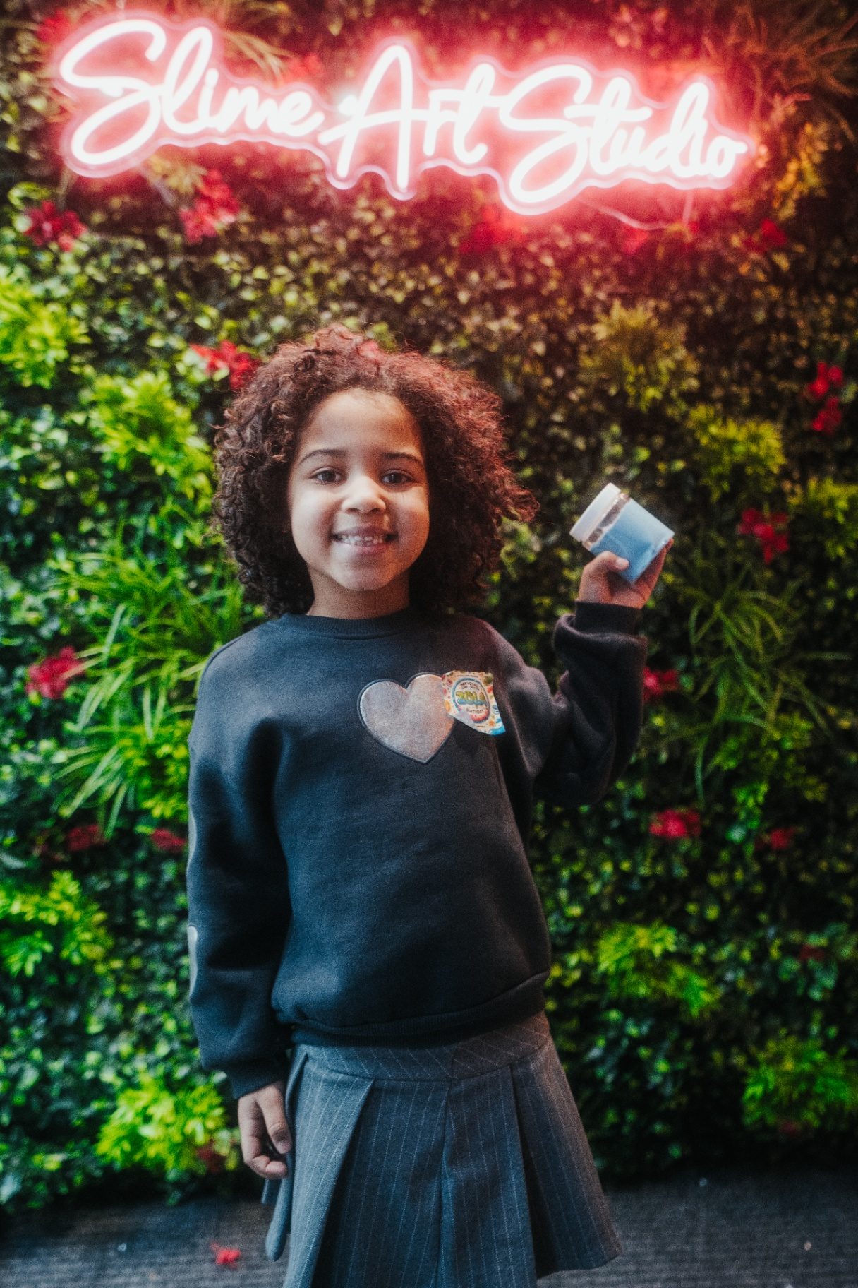 Atelier slime à Mons — enfant souriante devant le mur végétal et l'enseigne néon Slime Art Studio, tenant sa création